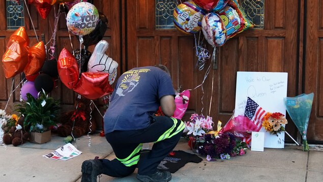 Un homme est agenouillé devant la porte d'une église, où l'on voit des ballons, des bouquets de fleurs et des messages écrits. Un homme est agenouillé devant la porte d'une église, où l'on voit des ballons, des bouquets de fleurs et des messages écrits.