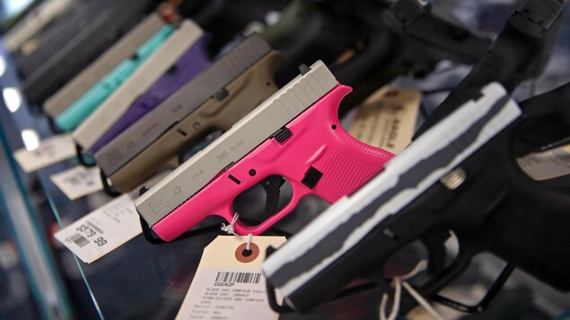 Des armes à feu de différentes couleurs sont sur un présentoir dans un magasin. Celle de couleur rose est mis en évidence, car la mise au point est effectuée sur elle.