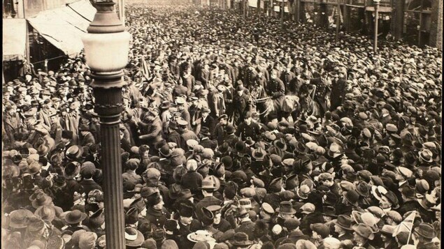 Une carte postale en noir et blanc montre une foule assemblée dans une rue.