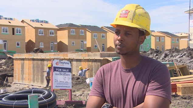 Un homme portant un casque de construction et ayant les bras croisés se tient devant le chantier de construction d'une maison dans un quartier résidentiel. Un homme portant un casque de construction et ayant les bras croisés se tient devant le chantier de construction d'une maison dans un quartier résidentiel.