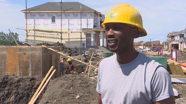Un homme portant un chapeau de construction sourit devant un chantier de construction où s'affairent deux autres hommes. Un homme portant un chapeau de construction sourit devant un chantier de construction où s'affairent deux autres hommes.