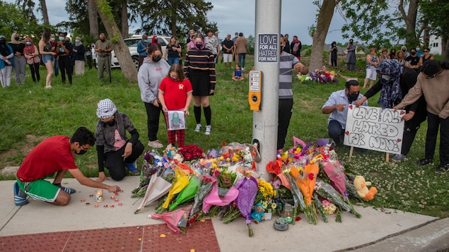 Ramos de flores junto a un poste con gente reunida en señal de duelo.