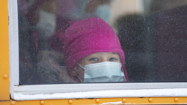 Une enfant coiffée d'une tuque rose et portant un masque bleu sur le visage est assise dans un autobus scolaire et regarde par la fenêtre.