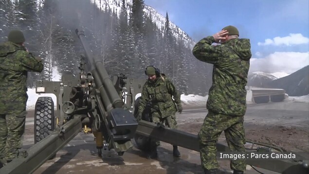Trois militaires, se bouchant les oreilles, sont à côté d'un canon d'où vient d'être lancé un projectileen direction de la montagne.