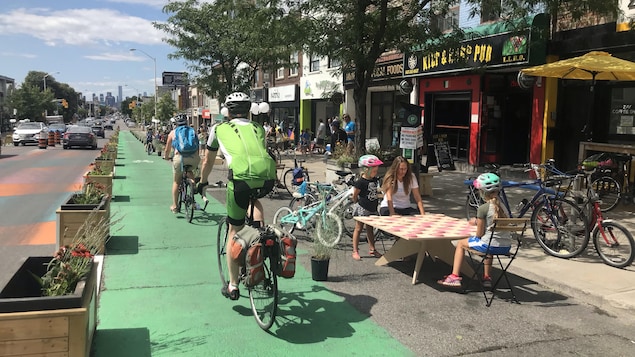Des cyclistes sur une piste cyclable avec des enfants qui jouent à côté sur un jeu de dames gégants avec un parent.
