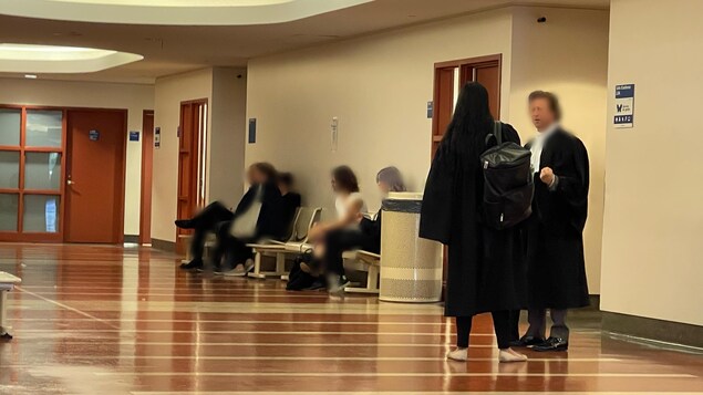 Deux avocats discutent dans le couloir d'un palais de justice où des visiteurs sont assis. 