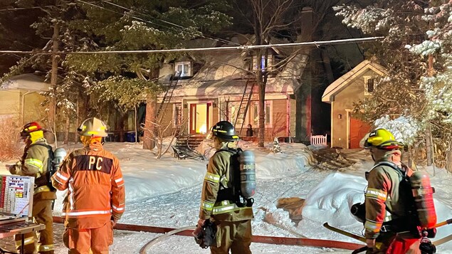 Quatre pompiers devant une résidence où des flammes sont visibles.