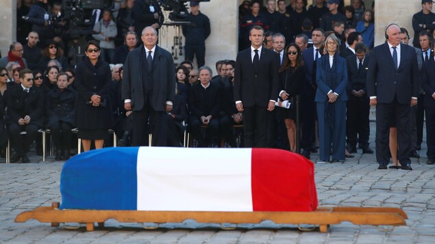 Un cercueil déposé sur un brancard derrière lequel se tiennent debout le président Macron et des dignitaires.