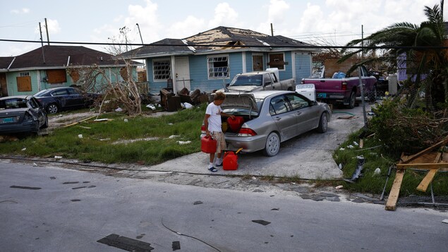 Un homme sort les bidons du coffre d'une voiture, dans un voisinage dont les maisons sont endommagées.
