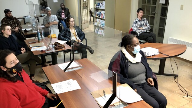 Les participants sont tous assis à des tables séparées par des plexiglas et écoutent un des responsables du programme.