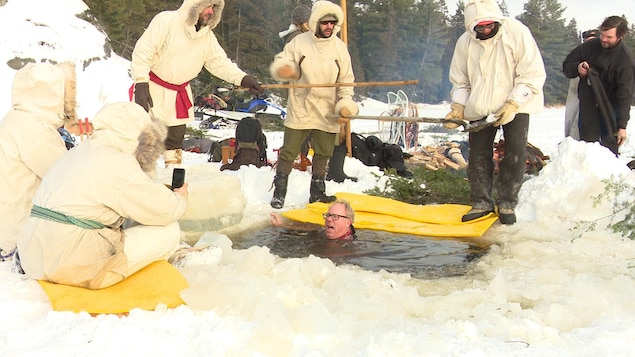 Au milieu d'un trou dans la glace, un homme est dans l'eau jusqu'au cou. À ses côtés, d'autres campeurs bien emmitouflés dans des anoraks se préparent à lui tendre des bâtons pour l'aider à sortir. 