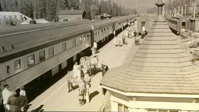 Sur cette photo en sépia, des passagers se tiennent sur une plateforme de train. Un train composé de nouveaux wagons en métal y est arrêté. Sur cette photo en sépia, des passagers se tiennent sur une plateforme de train. Un train composé de nouveaux wagons en métal y est arrêté.