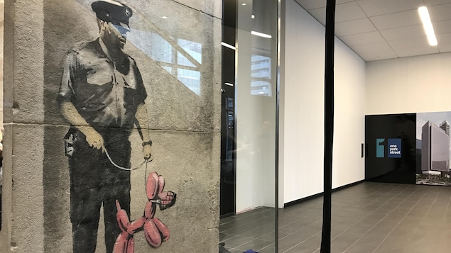 L'oeuvre de Banksy est au premier plan et derrière un plexiglas; au fond, une affiche du One York Street. L'oeuvre de Banksy est au premier plan et derrière un plexiglas; au fond, une affiche du One York Street.