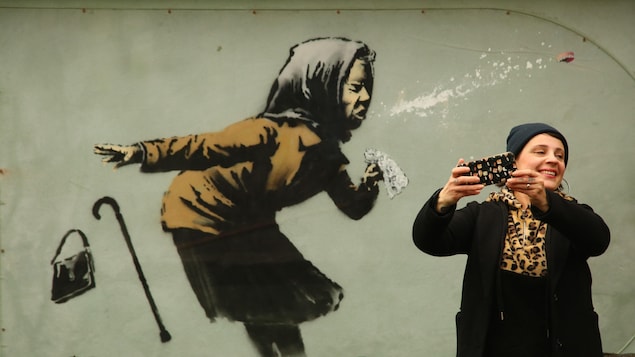 Une femme se prend en photo devant une murale de l'artiste de rue britannique Banksy illustrant une personne âgée qui éternue.