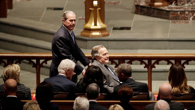 George W. Bush pousse le fauteuil roulant de son père; les cinq autres son assis et vus de dos. George W. Bush pousse le fauteuil roulant de son père; les cinq autres son assis et vus de dos.