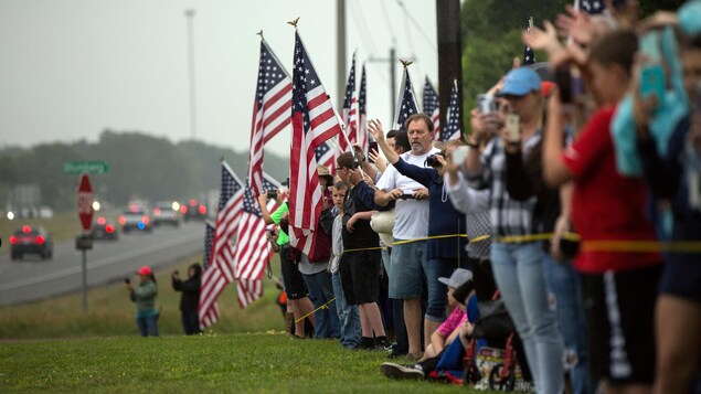 Une foule est assemblée le long d'un ruban jaune; plusieurs drapeaux des États-Unis sont visibles. Une foule est assemblée le long d'un ruban jaune; plusieurs drapeaux des États-Unis sont visibles.