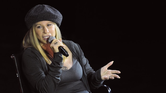 La chanteuse Barbra Streisand, toute vêtue de noir, parle dans un microphone lors d'une conférence.