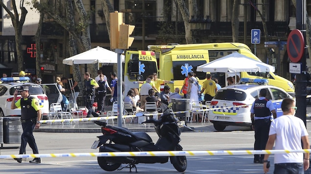 La police a établi un périmètre de sécurité dans le secteur la Rambla, à Barcelone. On y voit une ambulance et deux voitures de police, ainsi que plusieurs personnes derrière des cordons jaunes et blancs.