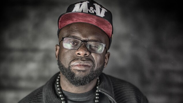 L'humoriste d'origine haïtienne porte casquette, lunettes et collier de barbe. Il fait une légère moue en regardant la caméra. L'humoriste d'origine haïtienne porte casquette, lunettes et collier de barbe. Il fait une légère moue en regardant la caméra.
