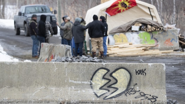 Au premier plan, un muret de béton sur lequel a été dessiné un cœur brisé. Au loin, un peu moins d'une dizaine de personnes sont rassemblées près de la barricade.