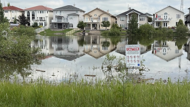 Une pancarte d'avertissement est engloutie sous les eaux, avec des maisons au loin.