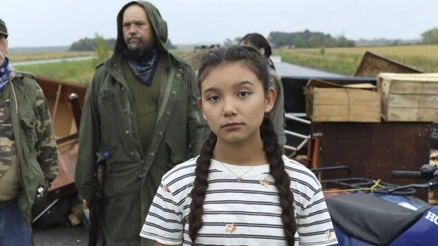 Une jeune fille regarde dans la caméra. Trois personnes vêtues d'imperméables sont en arrière-plan.