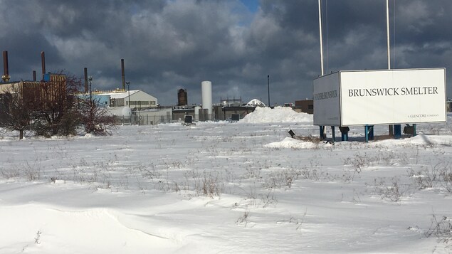 Un terrain enneigé au fond duquel on peut apercevoir une grande usine. 