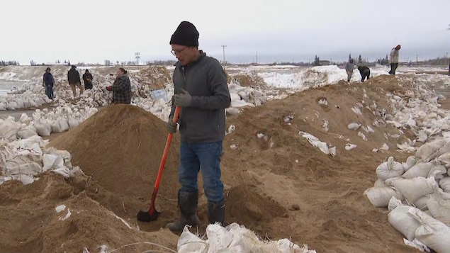 Un bénévole tient une pelle pour aménager une barrière constituée de sacs de sable à Peguis. D'autres bénévoles positionnent les sacs de sable afin de consolider les barrières.