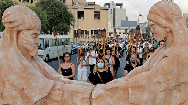 Des gens marchent en rang, torche à la main, derrière deux statues de terre.