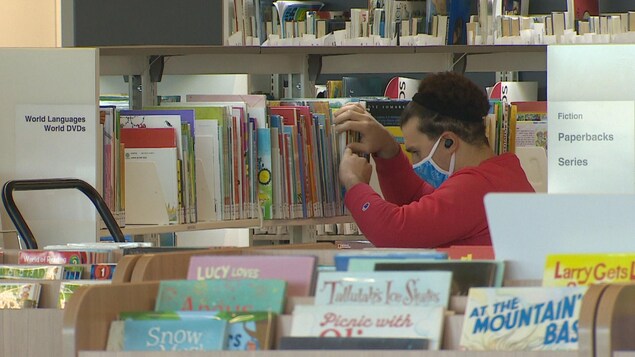 Une personne portant un masque cherche un livre rangés dans une étagère d'une bibliothèque