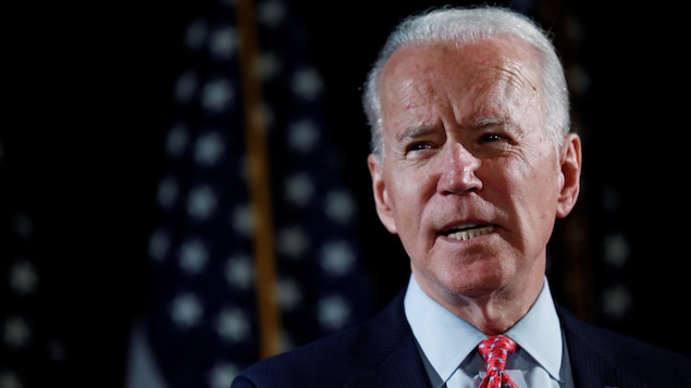Joe Biden regarde vers la droite, devant des drapeaux américains.