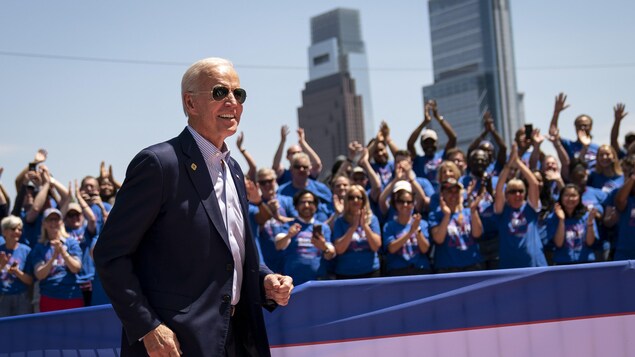 Un homme portant un complet et des lunettes de soleil sourit à une foule qui l'applaudit.
