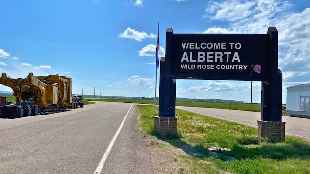 Un camion transporte une imposante pièce de machinerie jaune devant un panneau de bienvenue de la province de l’Alberta.