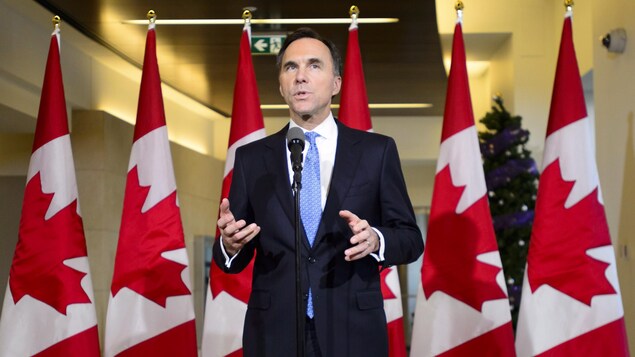 M. Morneau est debout au micro devant une série de drapeaux canadiens.