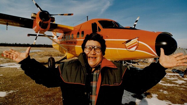 Un homme, les bras ouverts, sourit devant un petit avion orange.
