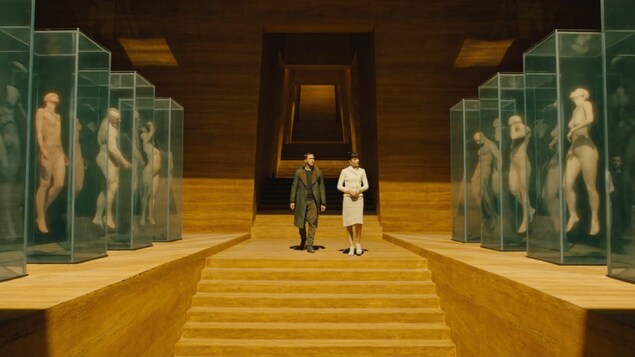 Deux personnages de Blade Runner 2049 déambulent dans une galerie de géants humanoïdes.