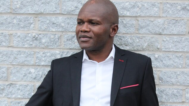 Portrait du juriste et écrivain d'origine congolaise Blaise Ndala, debout en veston et chemise blanche devant un mur de briques gris pâle.