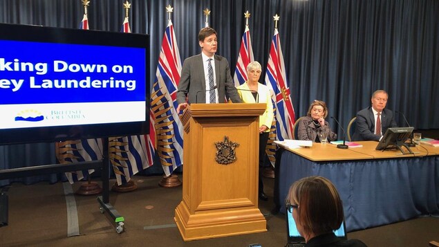Le procureur général de la province, David Eby, au podium. La ministre des Finances, Carole James, derrière. À leurs côtés, les auteurs des rapports, Maureen Maloney et Peter German.
