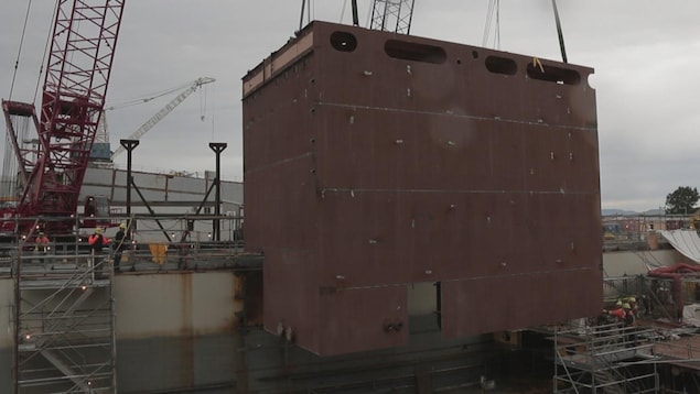 On voit un gros bloc brun, de côté, qui est hissé sur le chantier naval à l'aide de grues. On voit un gros bloc brun, de côté, qui est hissé sur le chantier naval à l'aide de grues.