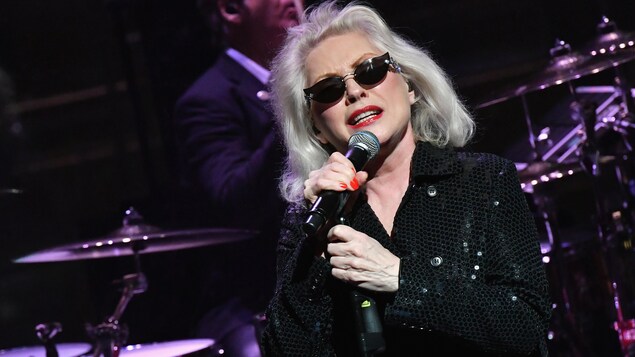 La chanteuse porte des lunettes fumées et chante au micro. La chanteuse porte des lunettes fumées et chante au micro.
