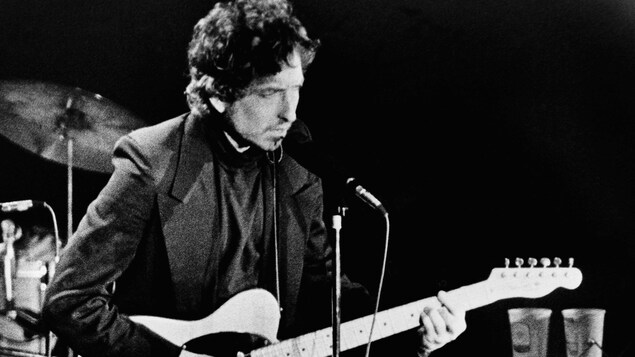 Bob Dylan chante dans un micro, en jouant de la guitare.