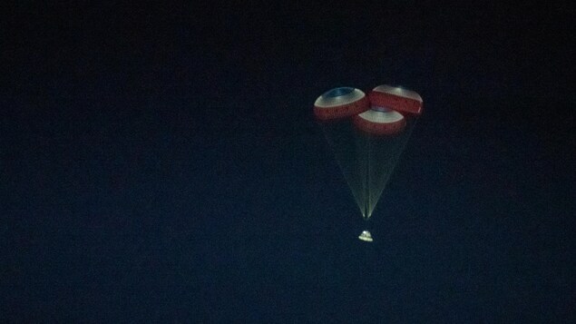 Une capsule spatiale descend dans le ciel, ralentie par trois grands parachutes.