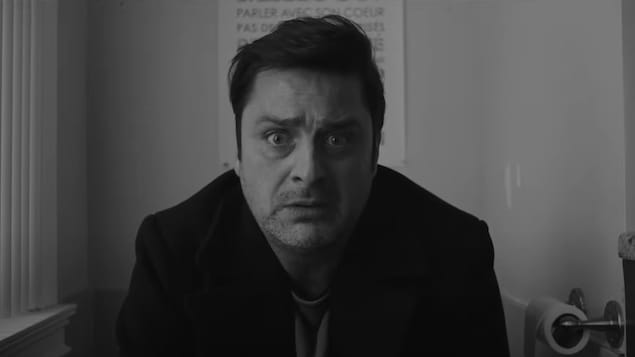 Un homme à l'air inquiet regarde droit devant lui, alors qu'il est assis dans une salle de bains, dans une image en noir et blanc de la série Bon matin Chuck.