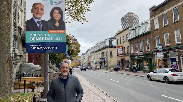 Un homme debout devant une affiche électorale accrochée à un poteau dans la rue.