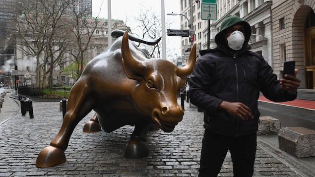 Une homme portant masque médical se photographie devant la statue à l'effigie d'un taureau devant la Bourse de New York.