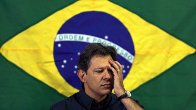 Le candidat de la gauche à la présidentielle brésilienne, Fernando Haddad, une main au visage devant un drapeau du Brésil.