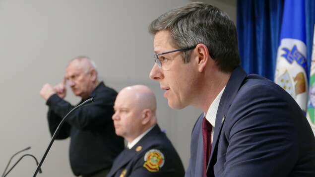 Brian Bowman est photographié de profil. Il apparaît à l'avant-plan de la photo. Plus loin se tient un représentant de la police de Winnipeg, et derrière on peut voir un homme qui fait l'interprétation en langage des signes.