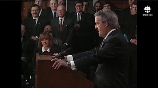 Brian Mulroney prononçant un discours au lutrin, sous le regard de sa femme Mila et de son entourage, en second plan
