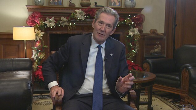 Brian Pallister, assis sur un fauteuil, en conversation, bouge la main. Derrière lui : des décorations de Noël autour du foyer dans son bureau.