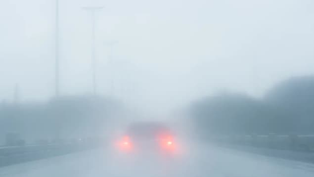 La lumière jaune orangé des phares d'une voiture apparaît derrière une épaisse couche de brouillard sur une route.